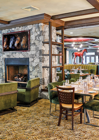 A restaurant interior with green chairs, a decorated carpet, stone fireplace, and horse sculptures in the background, table settings ready for guests.