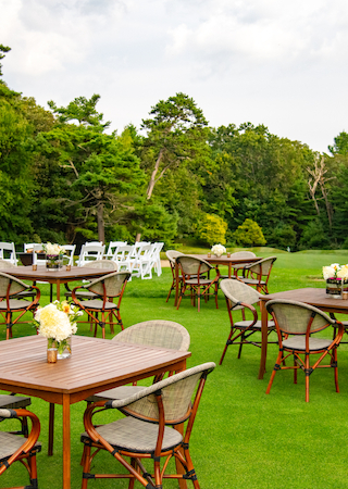 An outdoor event setup with wooden tables, cushioned chairs, and flower centerpieces on a grassy area surrounded by trees and additional seating in the background.