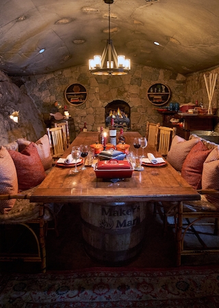 A cozy, rustic dining room with a wooden table, cushioned chairs, stone walls, and soft lighting, creating an inviting atmosphere for gatherings.