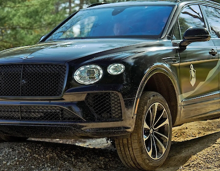 Black Bentley Bentayga driving on a dirt road surrounded by trees.