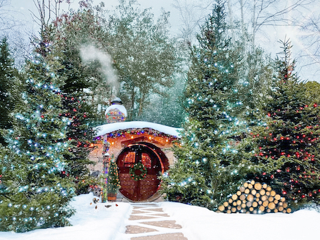 A cozy house with a round door, surrounded by snowy trees with colorful lights, smoke rising from the chimney, and stacked firewood.