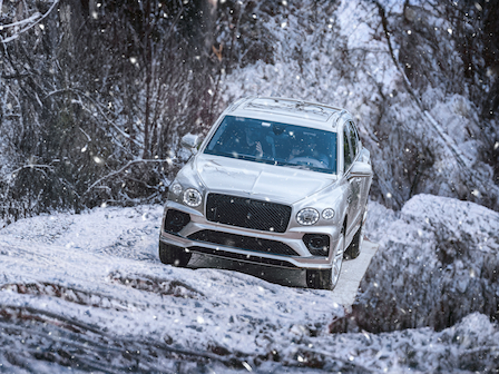 A silver SUV is driving on a snowy terrain with trees in the background, as snow falls gently around.