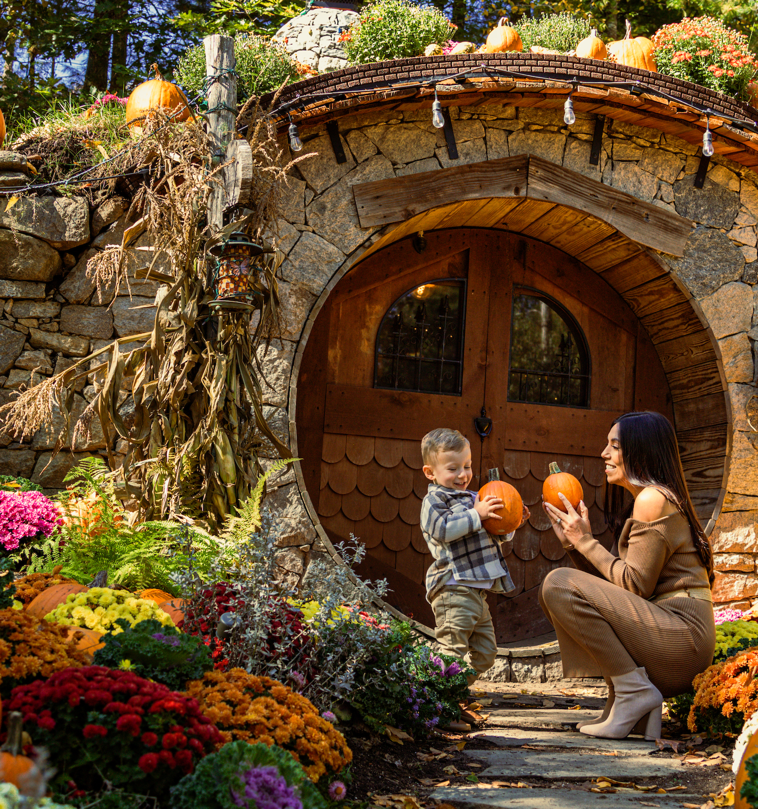 A woman and a child hold pumpkins outside a round, Hobbit-like door adorned with autumn decorations, surrounded by pumpkins and colorful flowers.