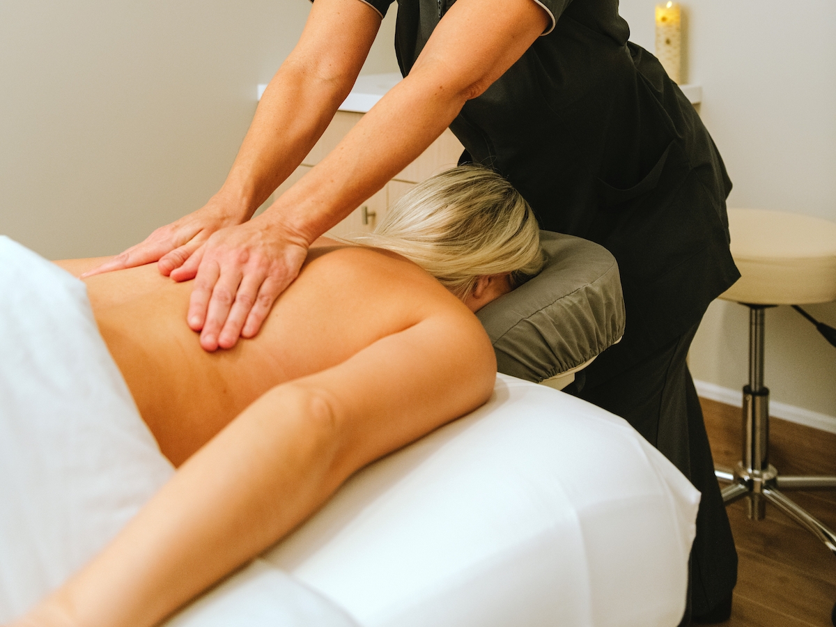 A person is receiving a back massage from a professional in a spa setting. The individual lies face down on a massage table covered with a towel.