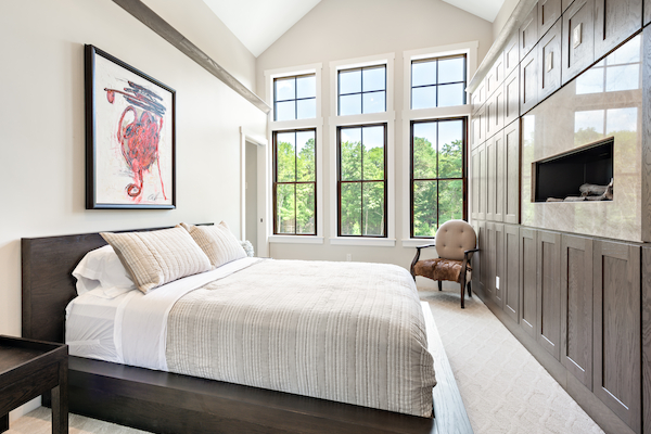 A modern bedroom with a large bed, artwork, tall windows, a chair, and built-in wooden cabinets with a recessed TV, filled with natural light.