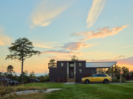 A modern house with a flat roof and large windows stands near a yellow car. The sky is painted with a picturesque sunset, and trees surround the property.