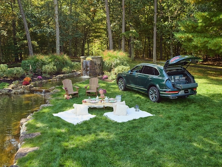 A car with its trunk open is parked by a serene pond, where a cozy picnic setup with blankets, chairs, and a table is arranged on the grass.