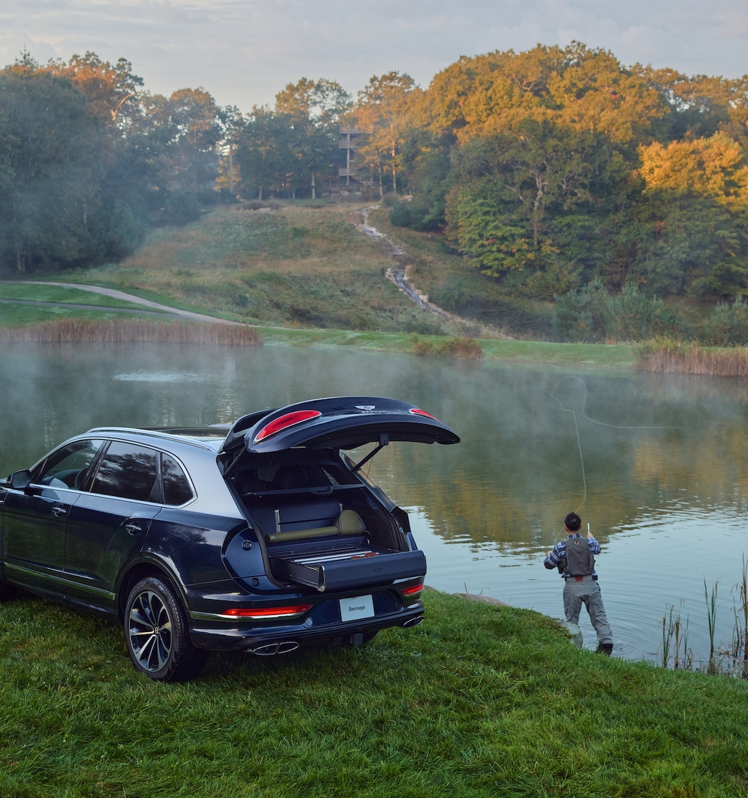 A parked SUV with an open trunk by a lake, a person is fishing nearby. The scene is set in a scenic, forested area with morning mist.