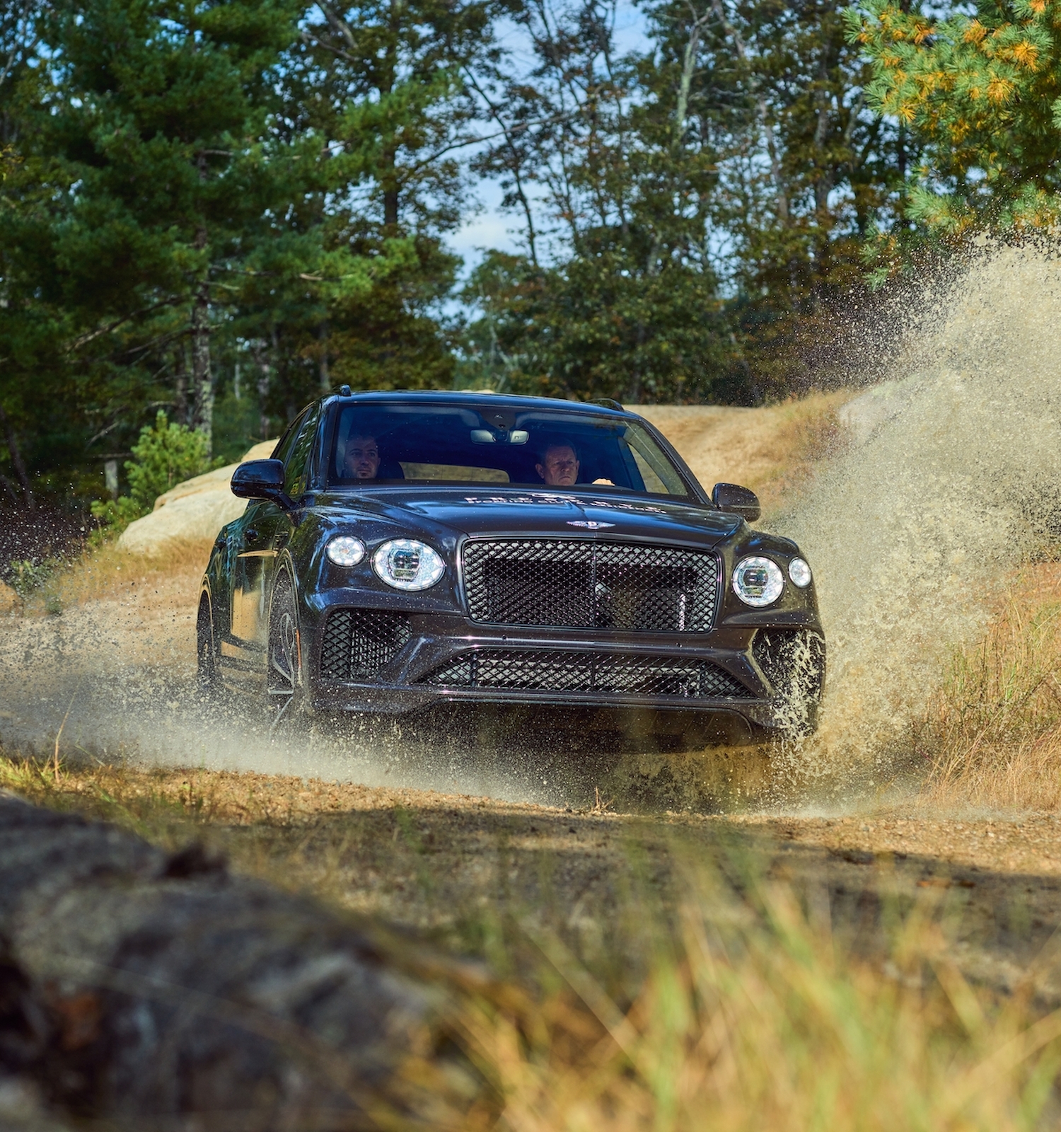 A black SUV is driving at high speed off-road on a dirt path, with dirt and dust flying up around it, surrounded by trees and vegetation.