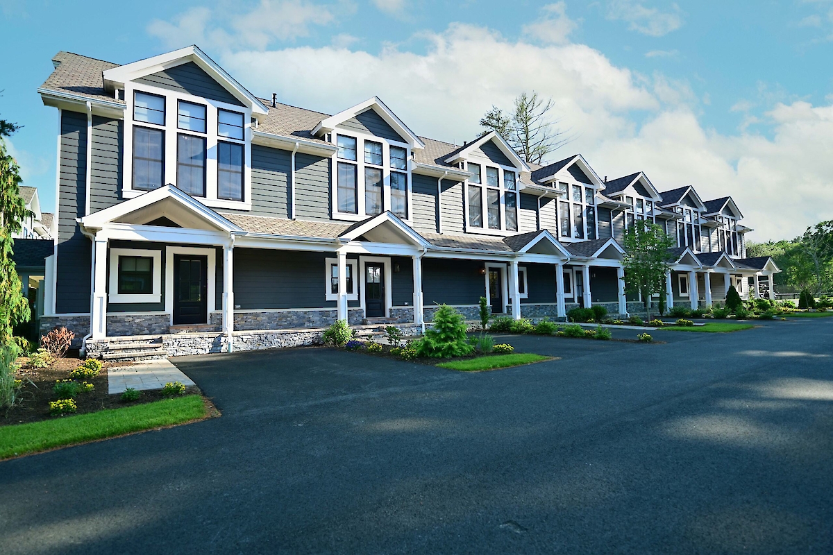 The image shows a row of modern two-story townhouses with porches and landscaped lawns, set against a clear sky.