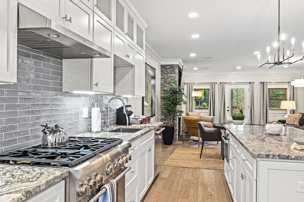 A modern kitchen with white cabinets, granite countertops, and stainless steel appliances opens to a cozy living area with large windows.