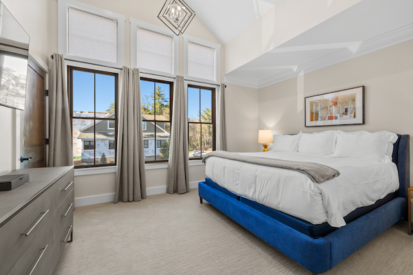 A well-lit bedroom with a large bed, blue bed frame, windows with curtains, dresser, framed wall art, and a lamp on a bedside table.