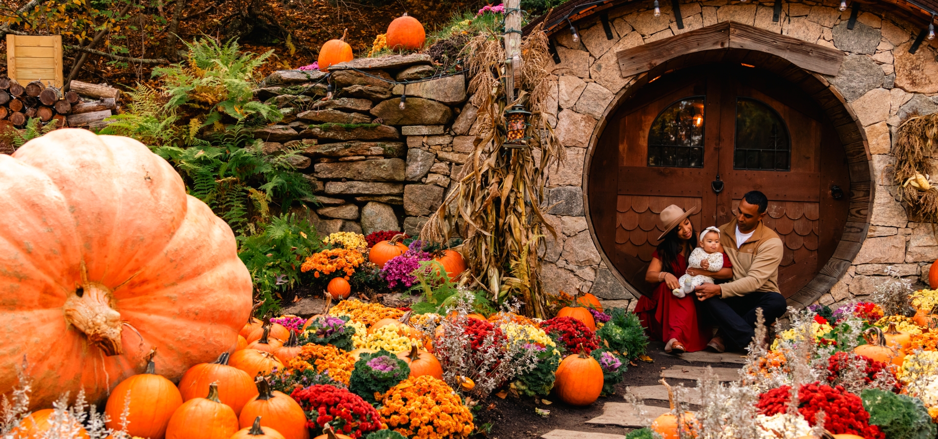 A family sits by a round doorway, surrounded by pumpkins and colorful flowers, creating a cozy autumn atmosphere.