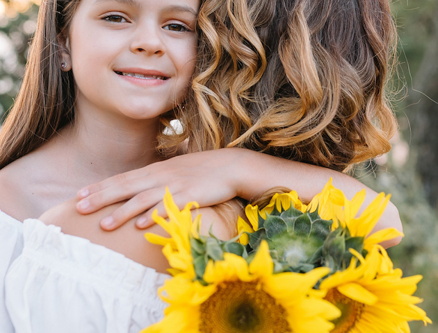 A young girl smiles, hugging another person holding bright yellow sunflowers. They stand outdoors, wearing white clothing.