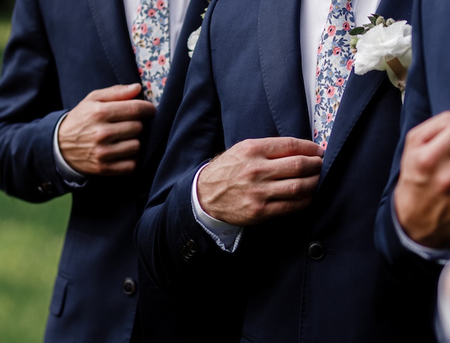 Two people in suits with floral ties, adjusting their jackets, possibly at a wedding, with one wearing a boutonniere on the lapel.