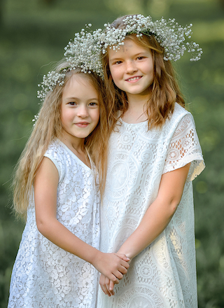 Two children in white dresses stand in a grassy area, wearing floral wreaths on their heads, holding hands, and smiling at the camera.