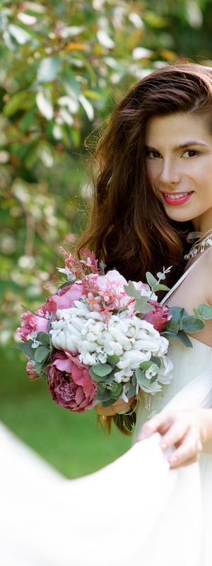 A woman in a white dress holding a bouquet of pink and white flowers, surrounded by greenery and smiling at the camera.