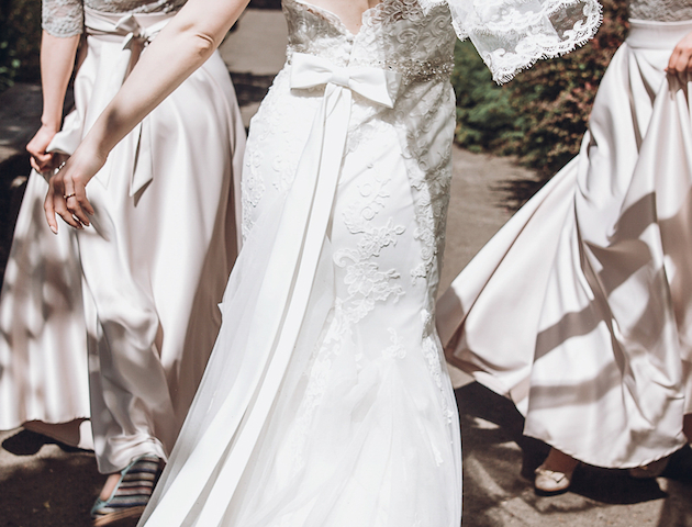 A bride in a white wedding dress and veil walks outdoors with two bridesmaids in light dresses, holding bouquets.