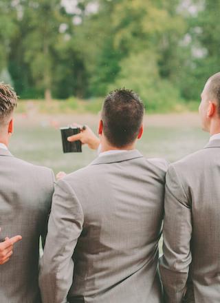 Three men in suits stand with their backs to the camera, one holding a flask, in an outdoor setting with greenery.