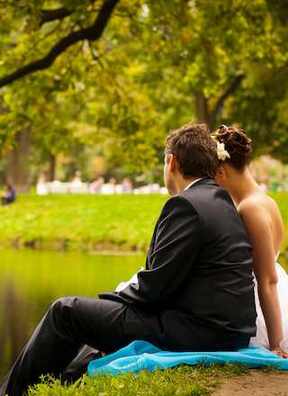 A couple, dressed formally, sits by a pond surrounded by greenery, enjoying a peaceful moment outdoors.