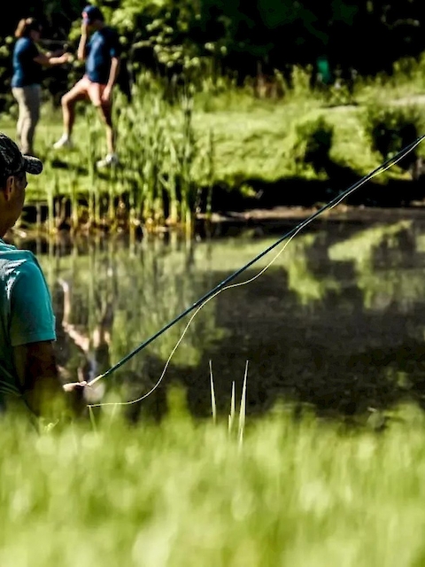 A person is fishing by a pond, surrounded by grass, with two more people walking in the background.