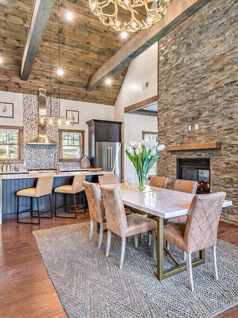 A modern open living space with dining area, kitchen, stone walls, wooden beams, chandelier, and stylish furniture decorates the room.