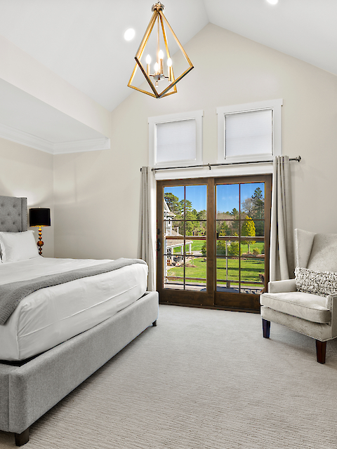 A spacious bedroom featuring a large bed, elegant armchair, chandelier, and glass doors leading to a scenic outdoor view.