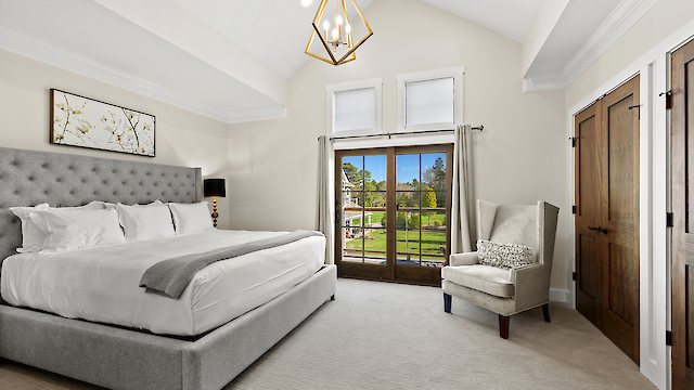 A spacious bedroom featuring a large bed, elegant armchair, chandelier, and glass doors leading to a scenic outdoor view.