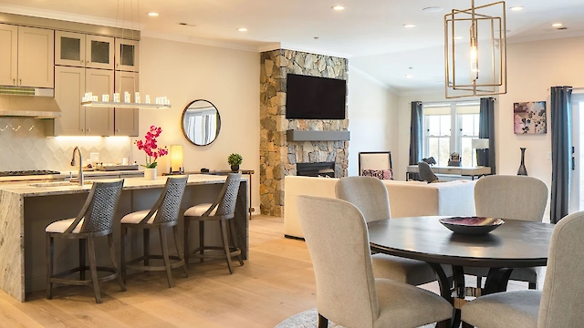 A modern open-plan living space with a kitchen, dining area, and cozy seating. Features neutral tones, warm lighting, a TV, and wall art.