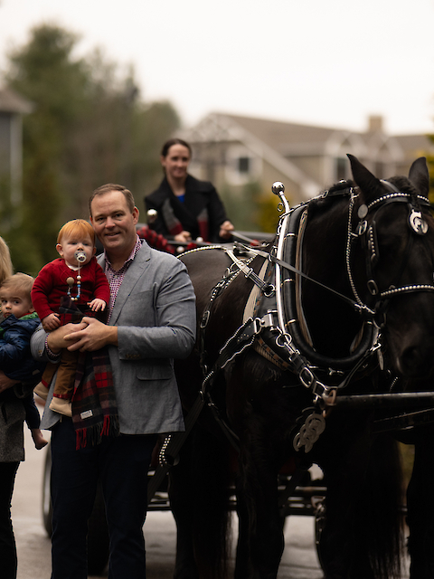 A group of people stands by a horse-drawn carriage with two dark horses on a residential street, smiling at the camera.