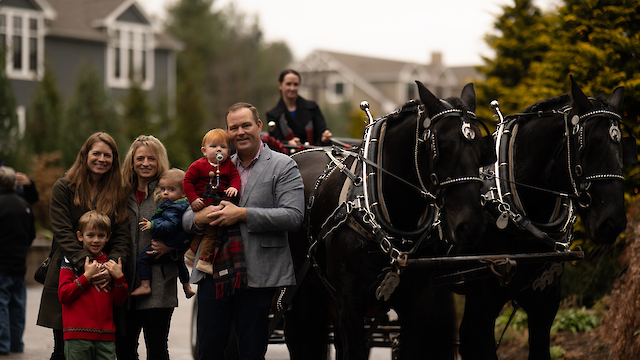 A group of people stands by a horse-drawn carriage with two dark horses on a residential street, smiling at the camera.