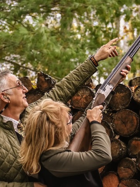 Two people outdoors, one aiming a shotgun upwards. Logs are stacked in the background.