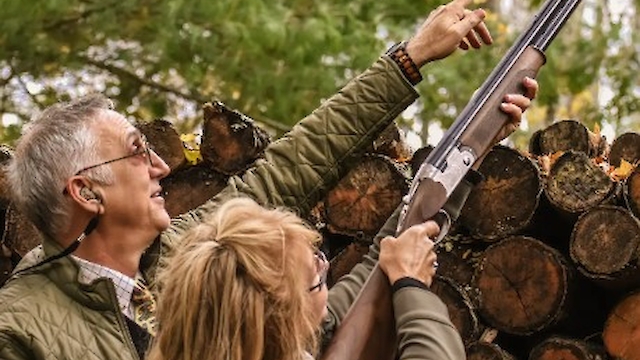 Two people outdoors, one aiming a shotgun upwards. Logs are stacked in the background.