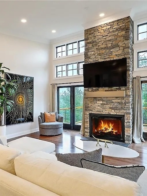 A modern living room with a white sofa, a large stone fireplace, a TV above it, and expansive windows opening to a view outside.