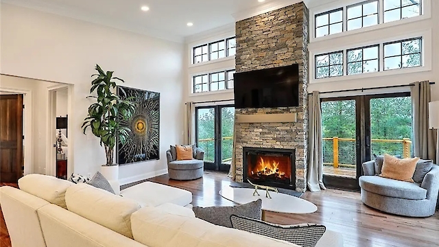 A modern living room with a white sofa, a large stone fireplace, a TV above it, and expansive windows opening to a view outside.