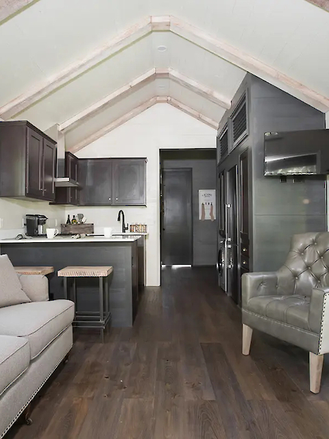 A modern tiny house interior with a kitchen, sofa, armchair, and large windows. Features wooden beams and sleek, neutral decor.