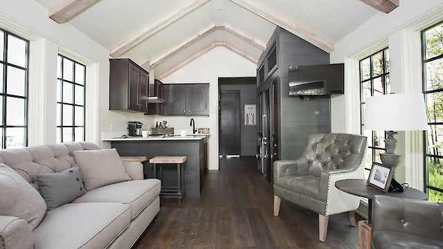 A modern tiny house interior with a kitchen, sofa, armchair, and large windows. Features wooden beams and sleek, neutral decor.