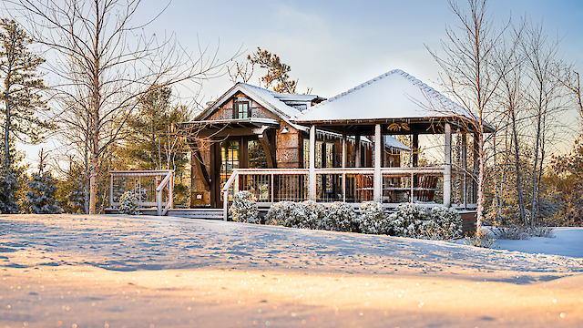 A cozy cabin with a wooden deck is covered in snow, surrounded by bare trees and a snowy landscape, under a clear sky.