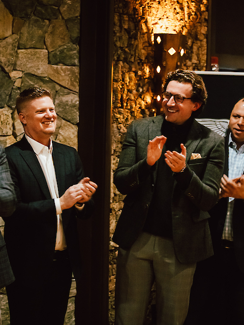 A group of four men in suits are standing and clapping in a warmly lit room.