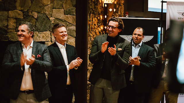 A group of four men in suits are standing and clapping in a warmly lit room.