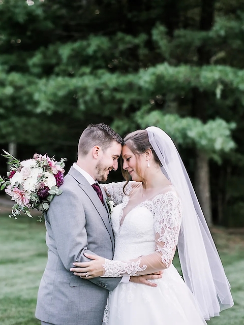 A bride and groom are embracing outdoors, with the bride holding a bouquet. They are surrounded by greenery.