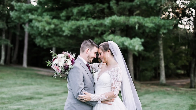 A bride and groom are embracing outdoors, with the bride holding a bouquet. They are surrounded by greenery.