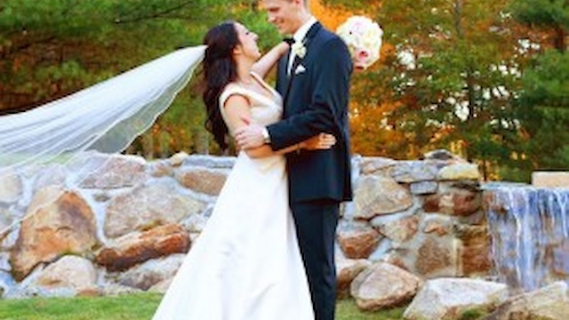 A couple in wedding attire poses outdoors on grass, near rocks and trees, with a long veil flowing behind the bride.