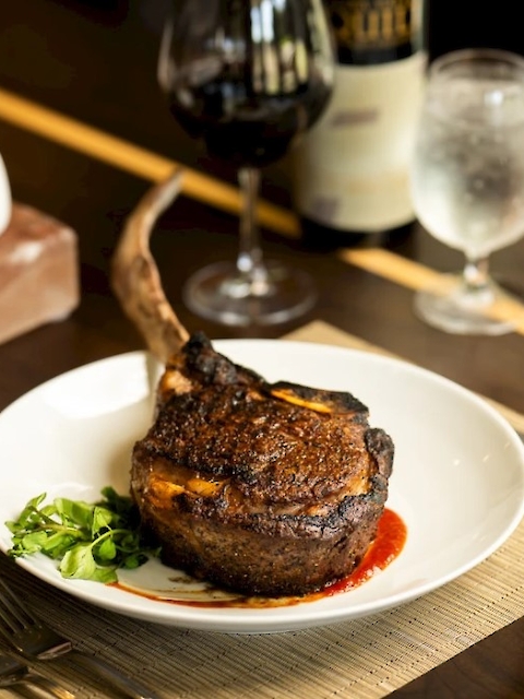 A cooked tomahawk steak is on a plate with a garnish, accompanied by a glass of red wine and a glass of water on a dining table.