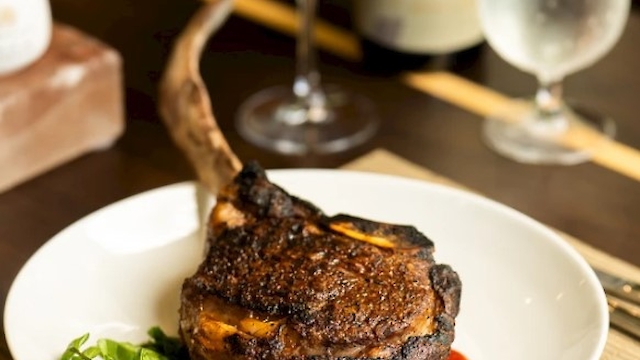 A cooked tomahawk steak is on a plate with a garnish, accompanied by a glass of red wine and a glass of water on a dining table.