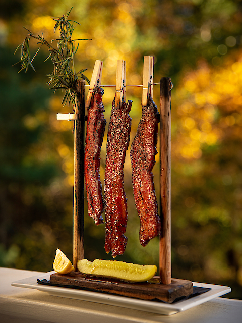 Three strips of cooked bacon hang from clothespins on a wooden frame, with a pickle and lemon wedge at the base.