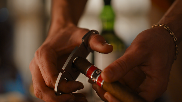 A person is using a cigar cutter to clip the end of a cigar. There’s a bottle in the blurred background.