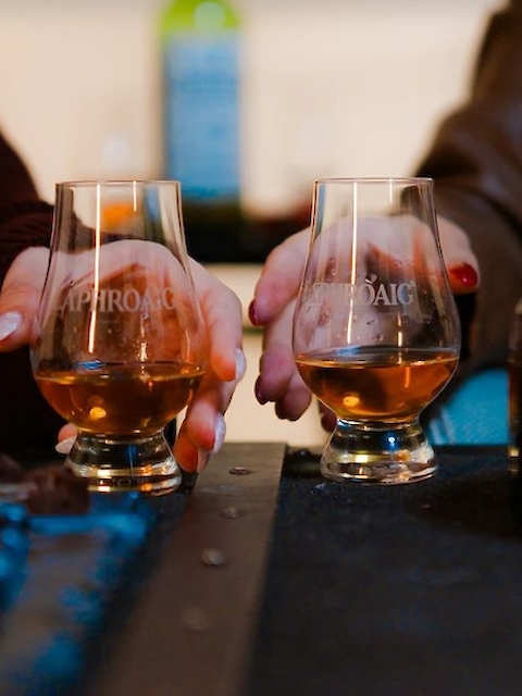 Two people are holding glasses of amber-colored liquid, possibly whiskey, with a blurred bottle in the background.