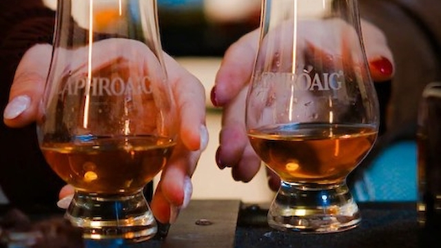 Two people are holding glasses of amber-colored liquid, possibly whiskey, with a blurred bottle in the background.