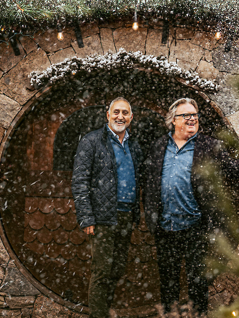 Two people stand in front of a round doorway framed by a stone wall, amidst falling snow and surrounded by trees with lights.